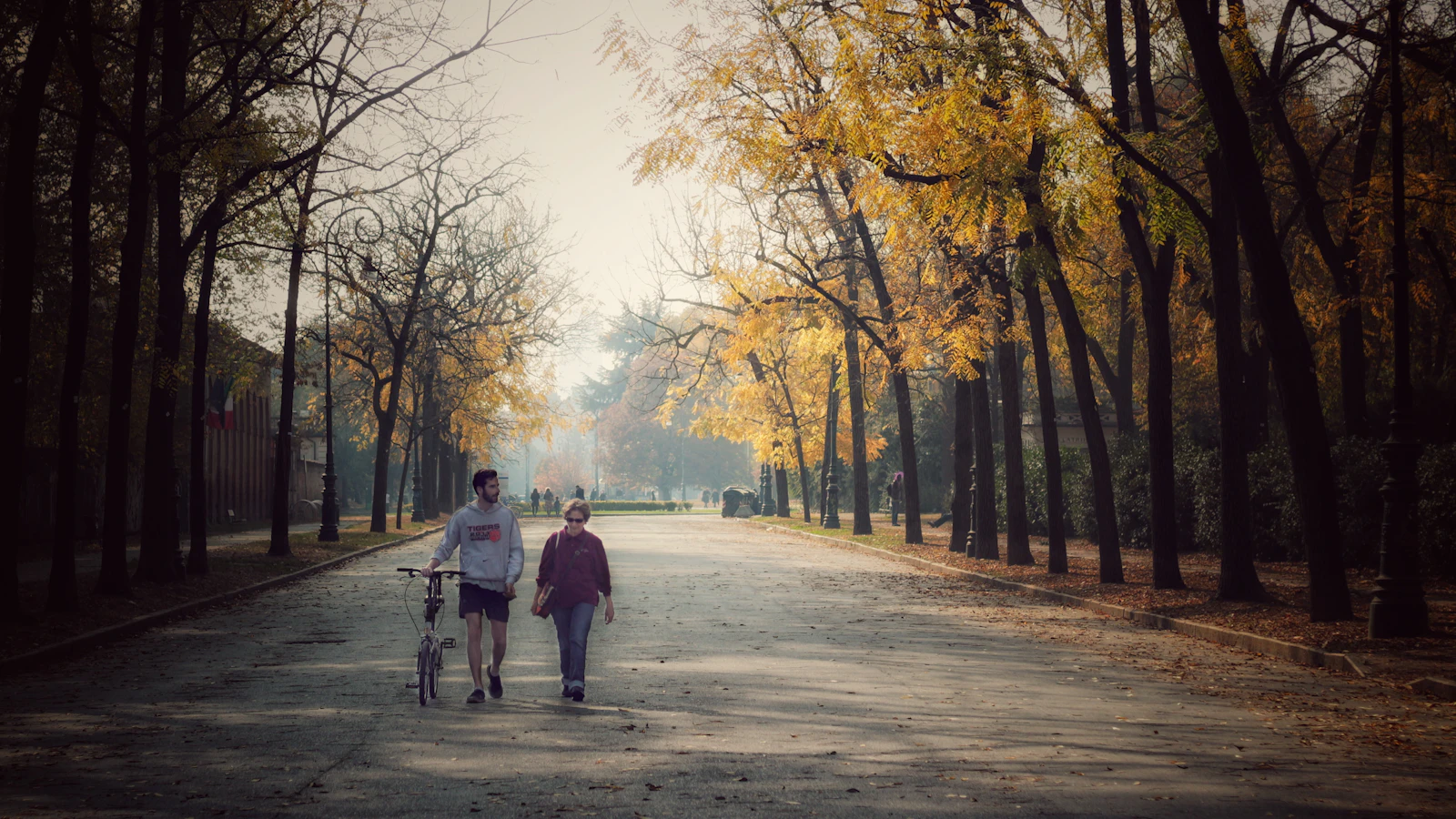 Een oudere vrouw wandelt met een jongere man over een laan met herfstbomen.