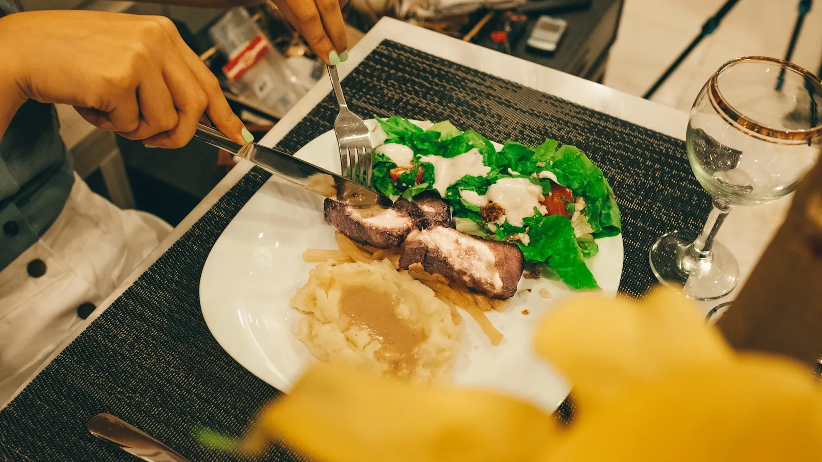 Iemand snijdt aan tafel een bord met vlees, aardappelpuree en salade.